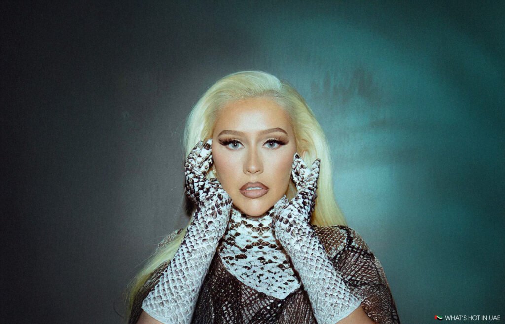 Person with long blonde hair wearing snake-patterned gloves, holding hands to face, against a teal background.