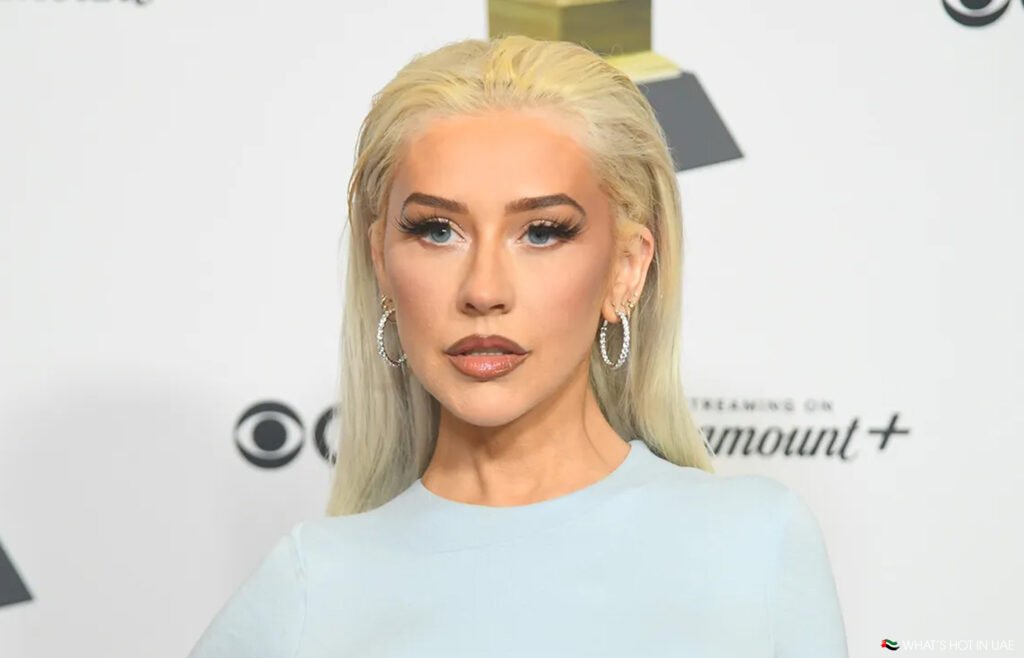 Christina Aguilera in Abu Dhabi with long blonde hair and wearing hoop earrings poses in front of a white background with logos.