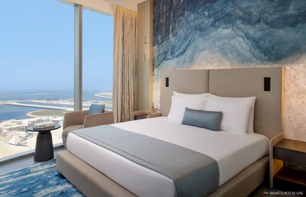 A modern hotel room with a large bed featuring white linens and a gray throw blanket, a small side table with a book, and an armchair by a floor-to-ceiling window overlooking a coastal view. The wall above the bed displays a blue abstract artwork.