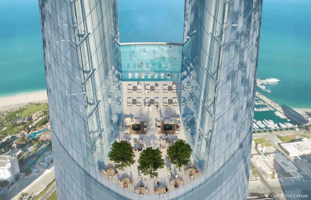 Aerial view of a Ciel Dubai Marina rooftop infinity pool on a high-rise building, surrounded by lounging areas and outdoor dining tables with umbrellas; the scene overlooks a marina and beach with a clear sky and ocean in the background.