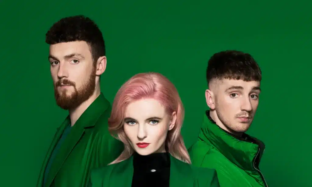 Clean Bandit posing against a green background, wearing green clothing; the central person has pink hair while the other two have dark hair.
