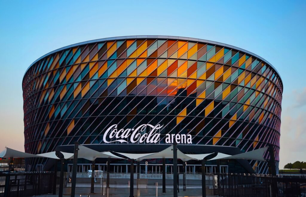 Megacampus Summit Dubai 2026: Exterior view of the Coca-Cola Arena featuring a colorful, diamond-patterned facade made of glass panels, illuminated by the sunset.