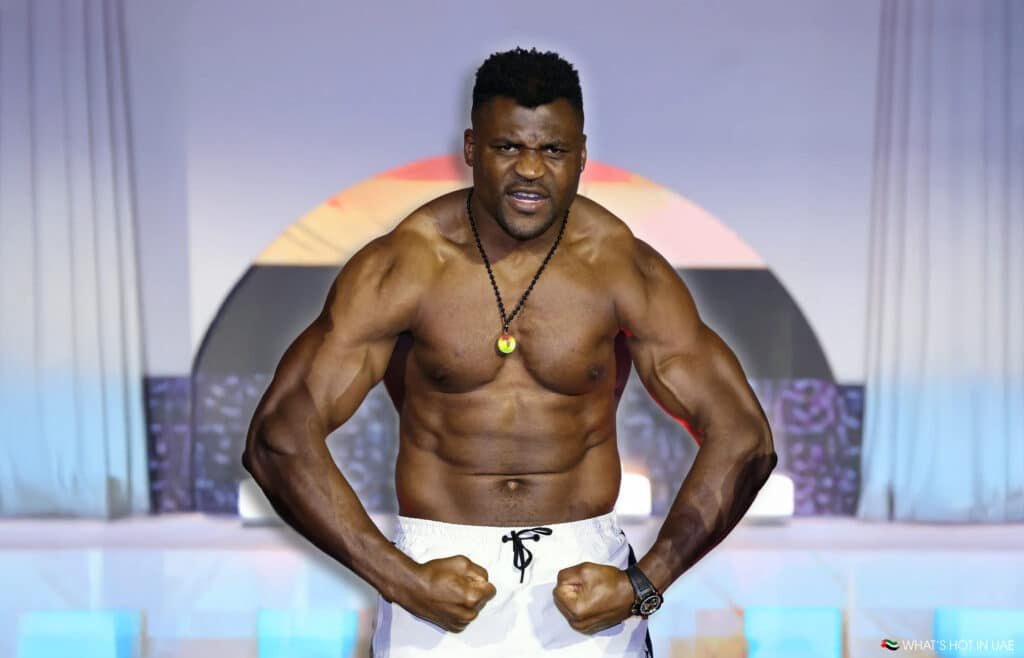 Francis Ngannou in white shorts flexing his arms, wearing a necklace and a watch, set against a colorful background.