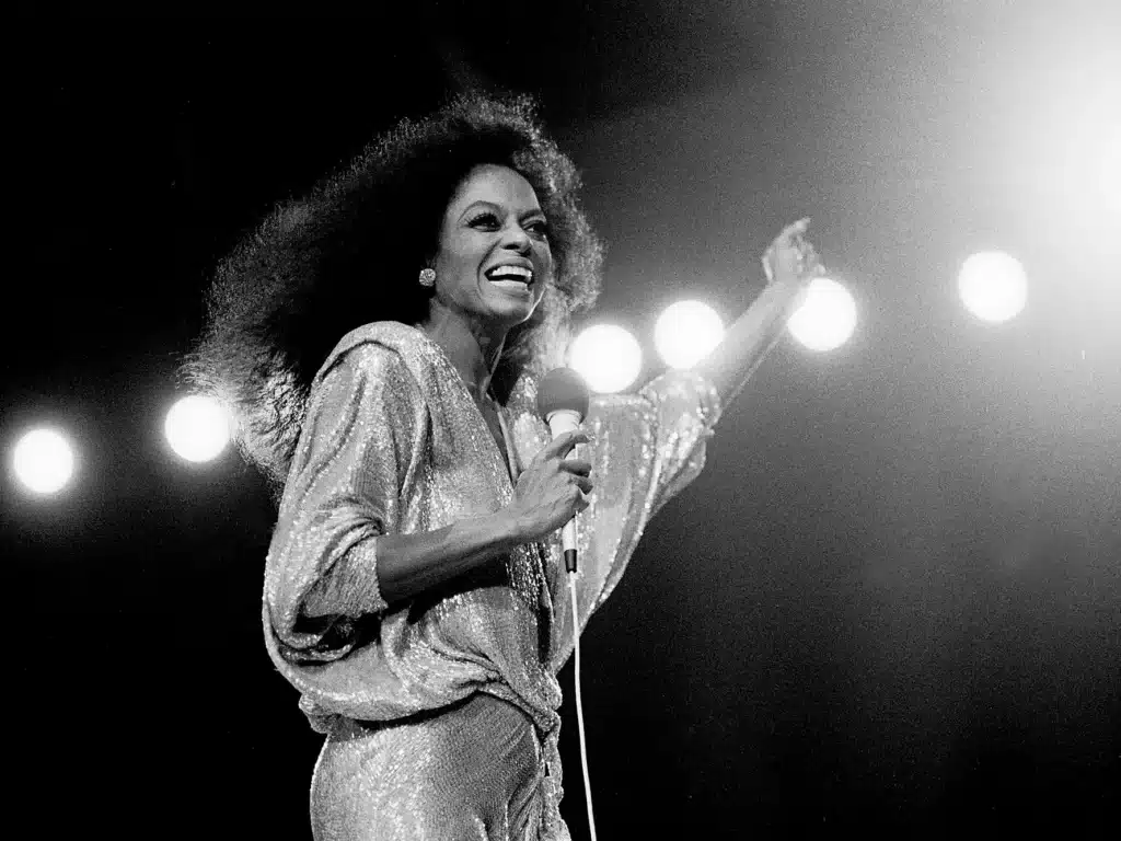 Diana Ross and Seal in a sparkling outfit singing on stage with a microphone, lit from behind by stage lights, smiling and raising one arm.