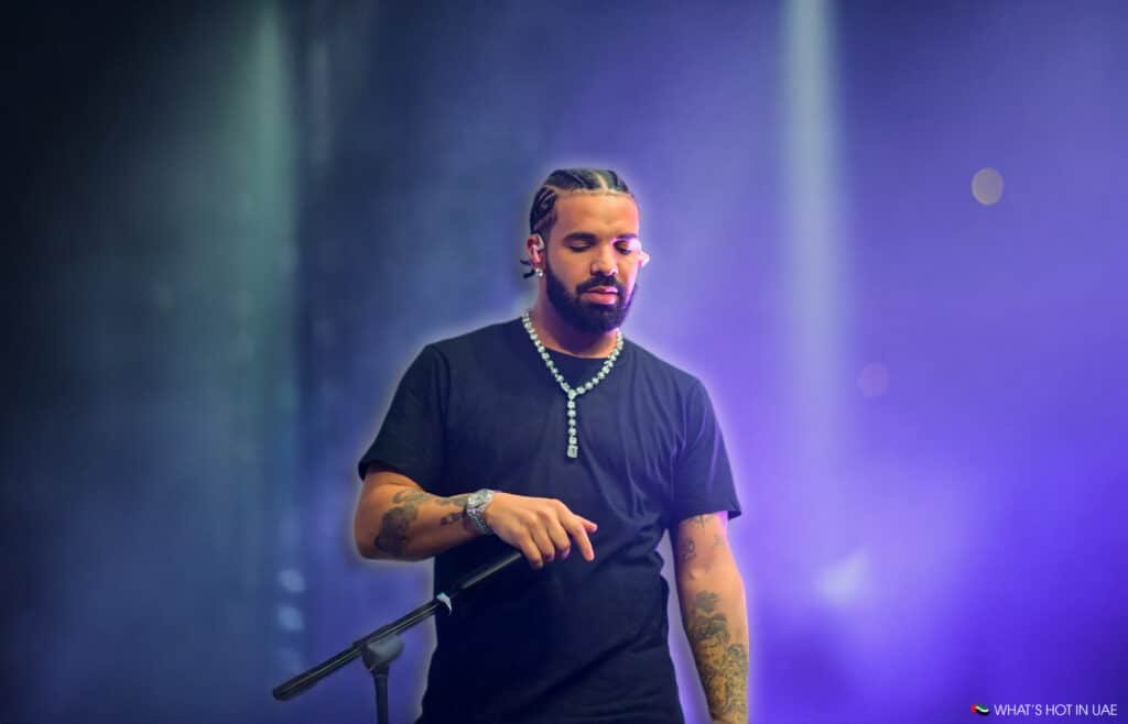 Drake with braided hair and a beard, wearing a black shirt and necklace, holding a microphone on a stage with dramatic lighting and a purple background.