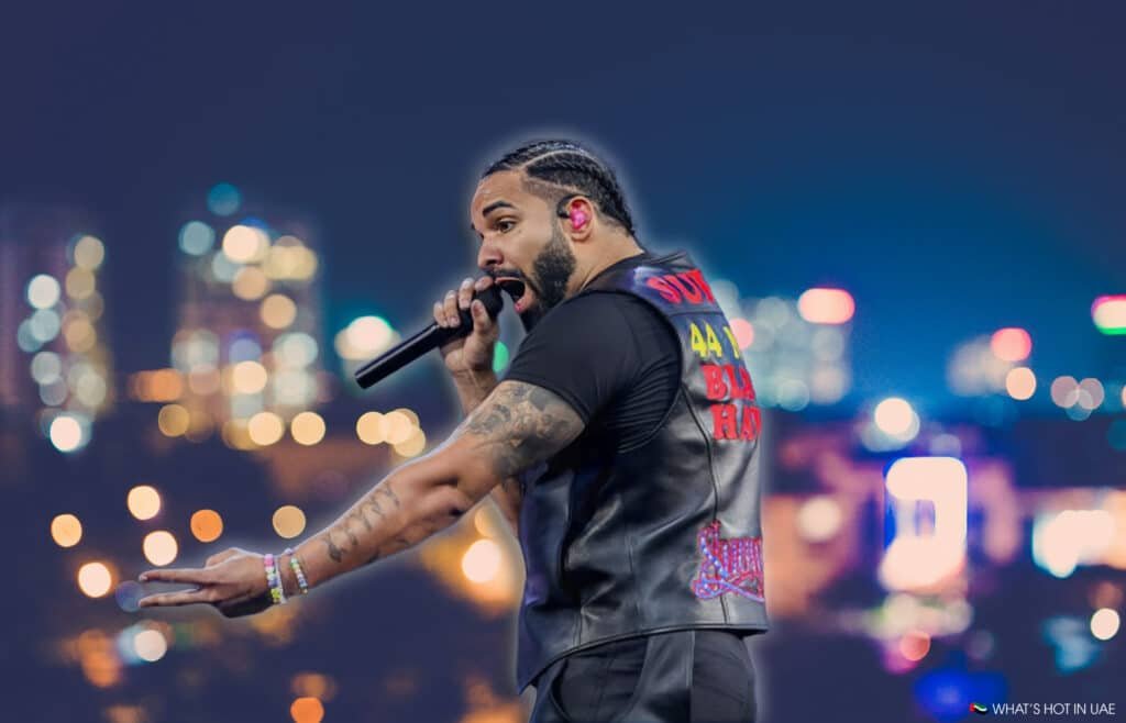 Drake performing on his World Tour 2026 stage, holding a microphone, with braided hair and wearing a black vest adorned with colorful text against a blurred city skyline backdrop at night.