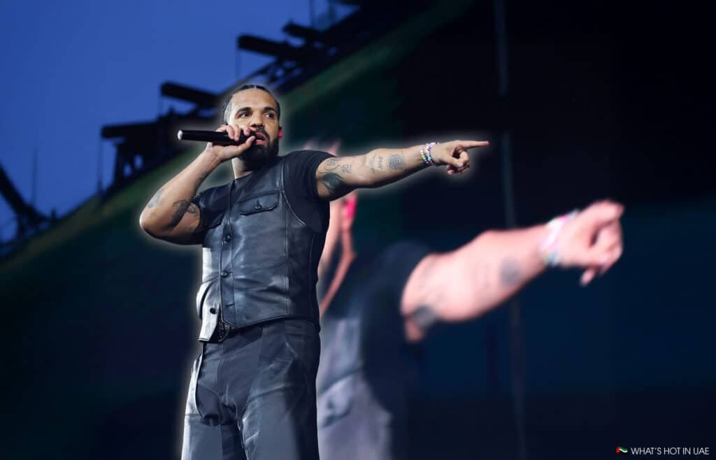 A performer on stage wearing a sleeveless leather vest, holding a microphone and pointing to the crowd, with a large screen displaying his image behind him.