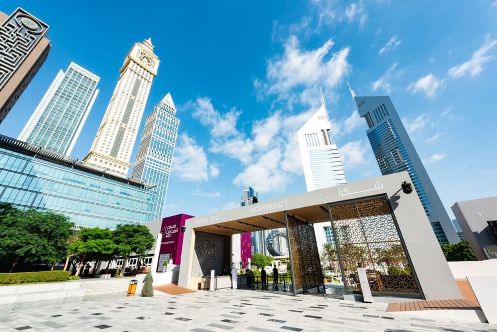 A modern cityscape featuring tall skyscrapers under a blue sky, with an entrance labeled 'Dubai Watch Week' in the foreground.