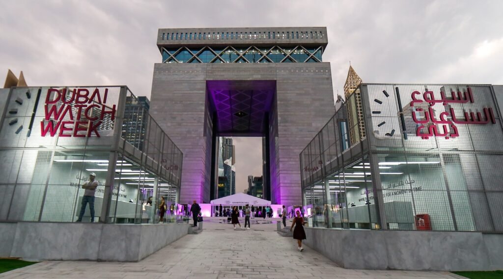 Entrance to Dubai Watch Week with signs and a large, modern stone archway illuminated in purple light, people are walking towards the exhibition hall.