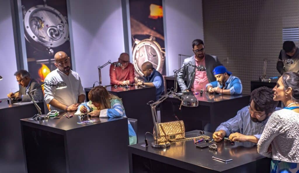A group of people working at individual desks with tools and lamps, possibly engaged in watchmaking or jewelry repair, within a well-lit room.