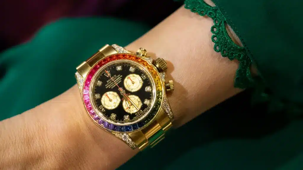 A gold wristwatch with a rainbow bezel and black dial, featuring three sub-dials and diamond hour markers, worn on a person dressed in a green outfit with lace trim.
