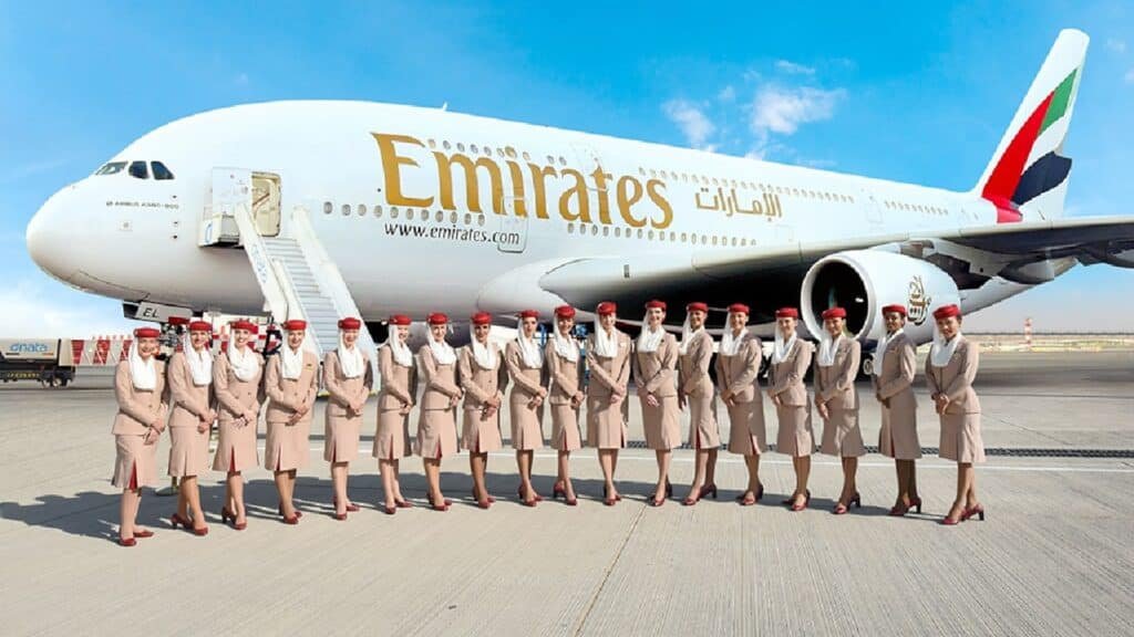 A group of Emirates flight attendants in uniform stand in front of an Emirates Airbus A380 parked on an airport tarmac.