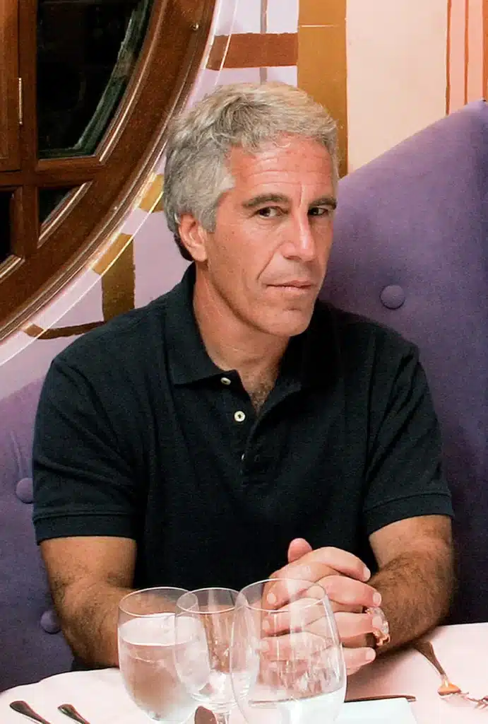 A man with gray hair and a black shirt sits at a table set with glasses and utensils, with a decorative wall and circular window in the background.