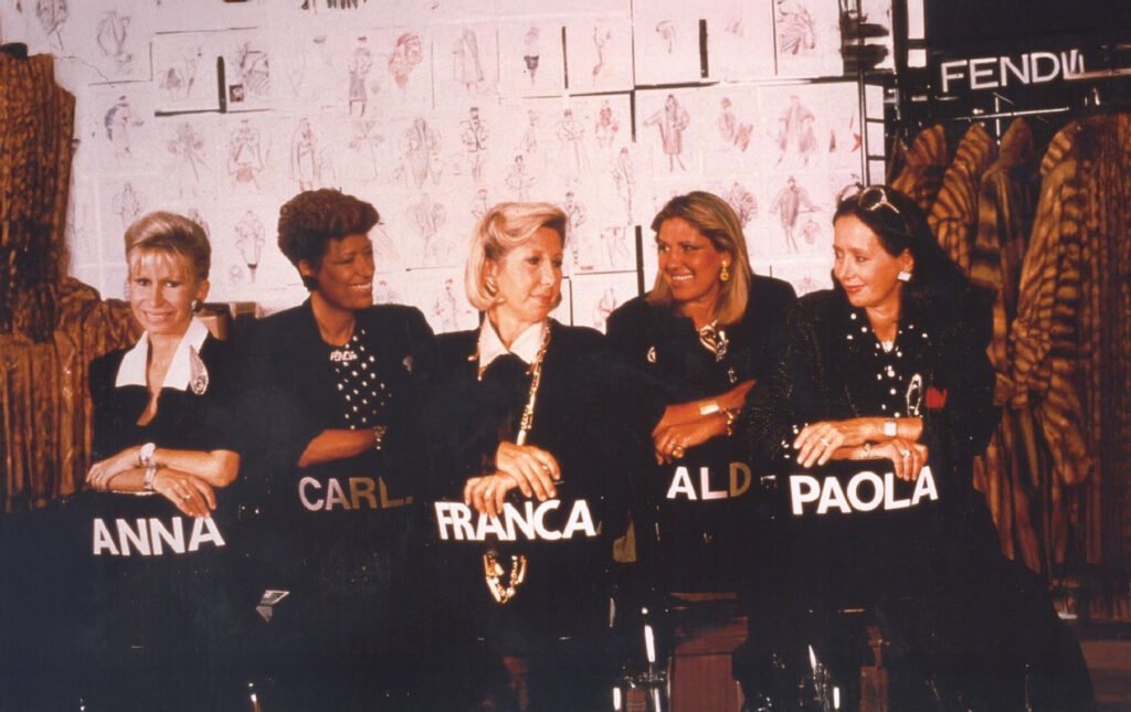 Five women sitting side by side, each with a chair labeled with their name: Anna, Carla, Franca, Alda, and Paola, against a backdrop of fashion sketches and coats labeled 'Fendi.'