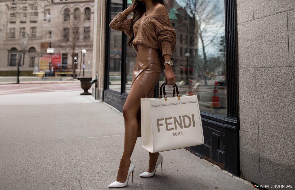 The History of Fendi: A woman wearing a brown off-shoulder sweater and a glossy tan skirt, holding a large white Fendi shopping bag, and walking in an urban setting with reflections in the glass window nearby.