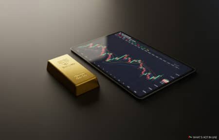 A gold bar labeled "GOLUSD" rests beside a tablet displaying a financial trading chart on a black surface.