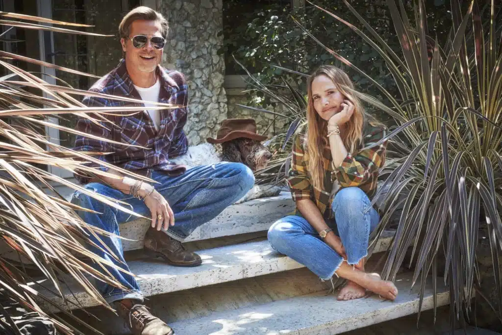 A man wearing sunglasses and a plaid shirt sits next to a woman with long hair, wearing a plaid shirt and jeans, on outdoor stone steps. A dog wearing a brown hat is lying between them. They are surrounded by greenery and large plants.