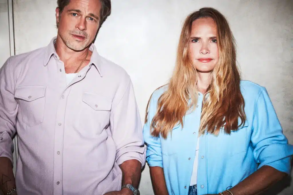 Brad Pitt's Luxury Fashion Brand in a light purple shirt and a woman in a blue shirt stand side by side against a light-colored wall, looking directly at the camera.
