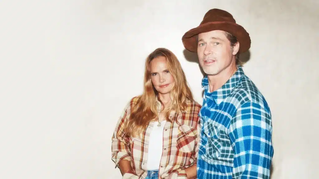 A woman with long blonde hair in a plaid shirt stands next to a man in a blue plaid shirt and brown hat against a plain background.