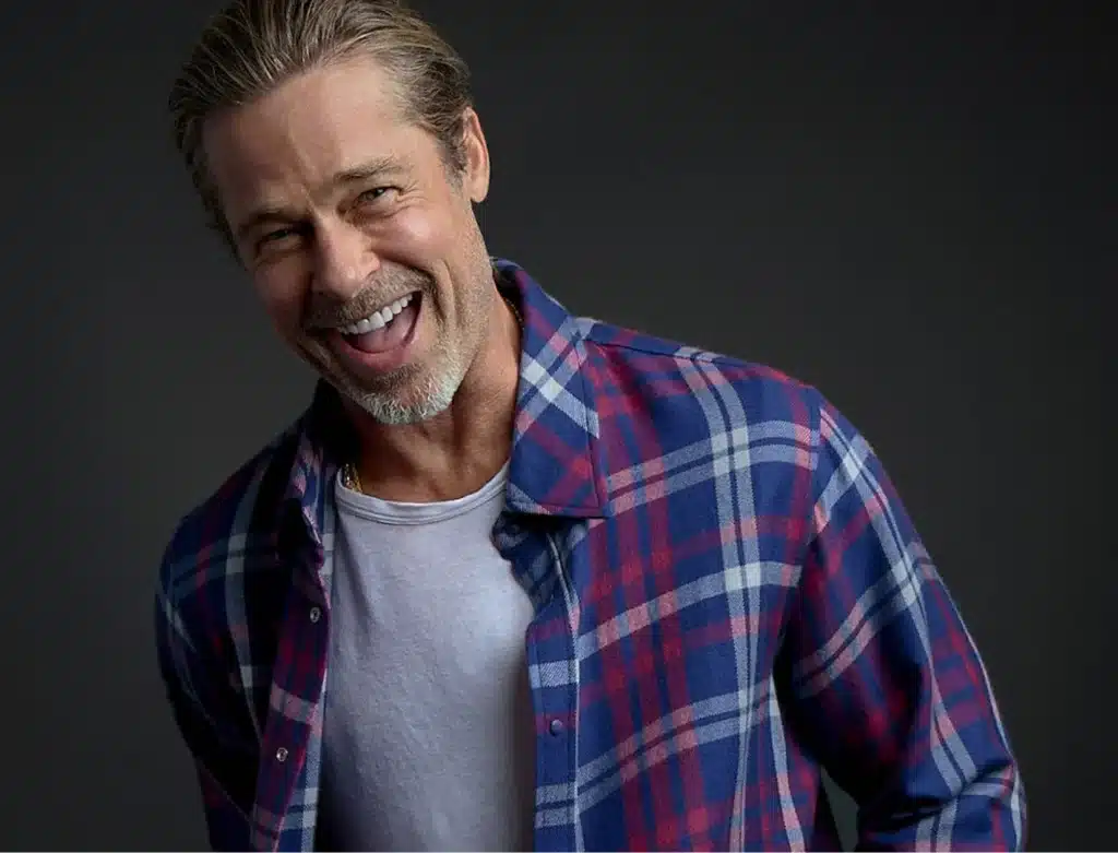 Brad Pitt's Luxury Fashion Brand smiling broadly, wearing a red and blue plaid flannel shirt over a white t-shirt against a dark background.