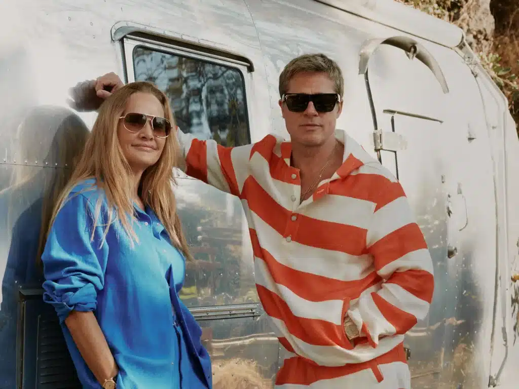 Two people wearing sunglasses stand leaning against a shiny metal surface; one wears a blue shirt, the other a red and white striped shirt.