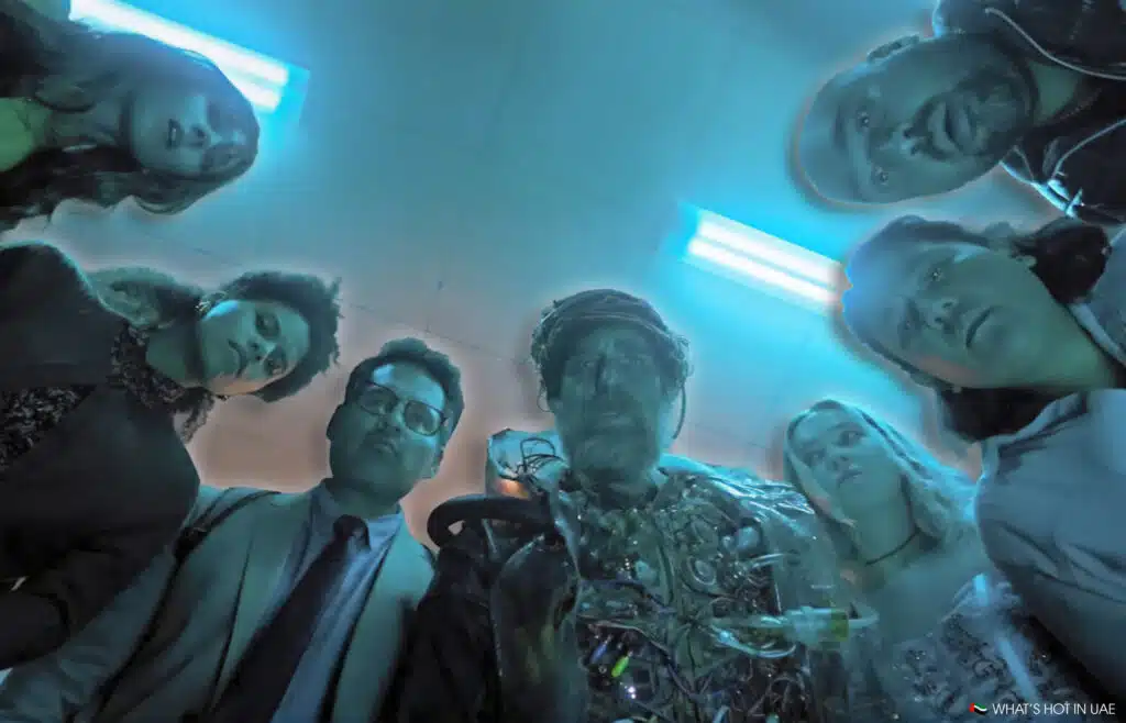 A group of people with curious expressions looking down, illuminated by a blue light, with one person in the center wearing a futuristic, gadget-laden costume.