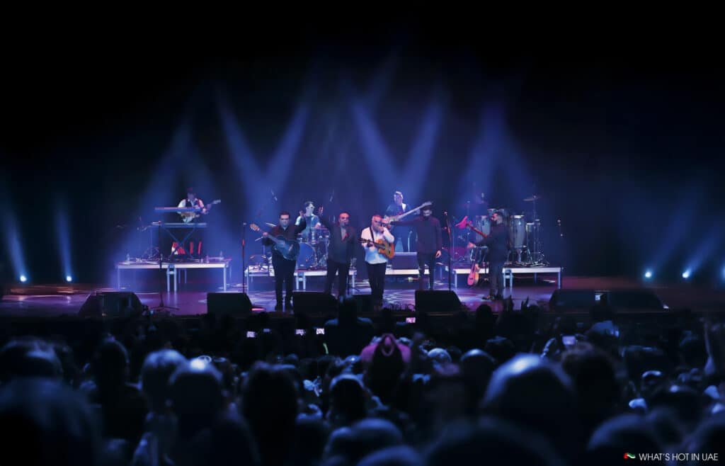 A band performing on stage with several musicians playing instruments under colorful, focused stage lights, while a large audience is visible in the foreground.