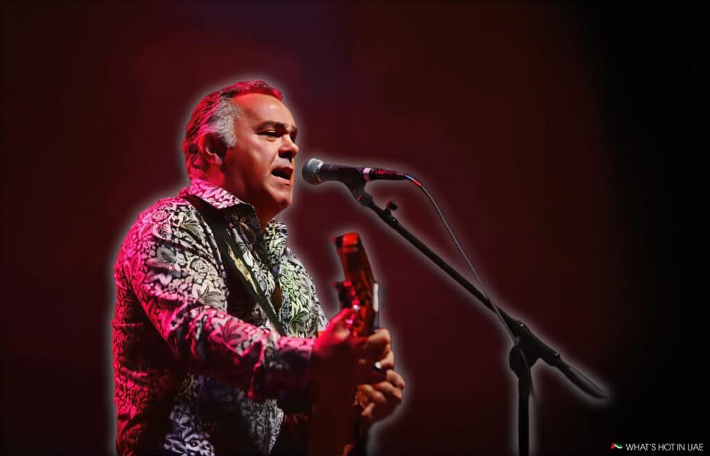 A performer singing and playing guitar on stage, illuminated by red lighting. The musician is wearing a patterned shirt and is positioned close to a microphone, with a focused expression.
