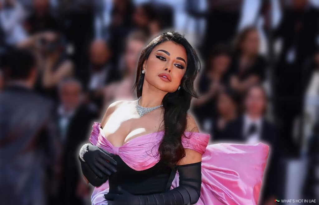 A woman wearing a glamorous pink dress with black gloves poses confidently on the red carpet, surrounded by a blurred crowd.