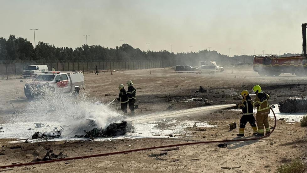 Indian Tejas Fighter Jet Crash. Firefighters in protective gear spray foam on a burning object in an open, dusty area, with a fire truck and other vehicles in the background.