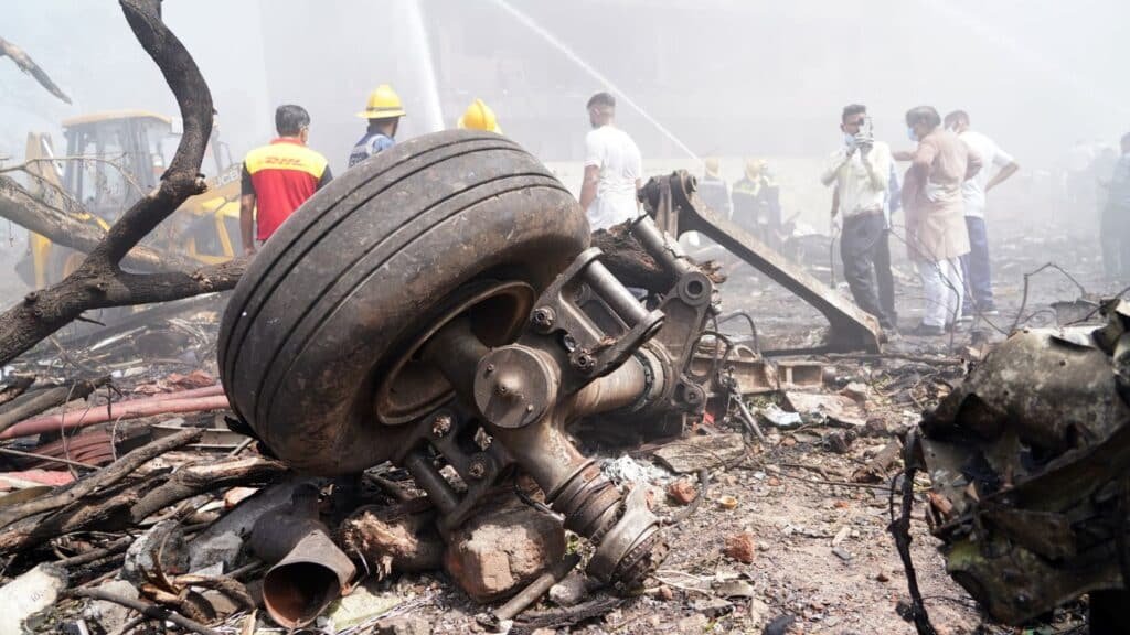 A crashed airplane wheel amidst debris with firefighters in the background working amidst smoke at the scene of an accident.