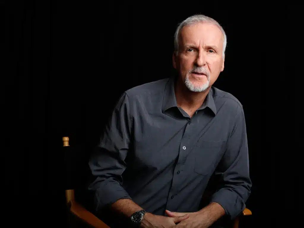 James Cameron with short white hair and a beard, wearing a dark grey button-up shirt, sitting against a black background.