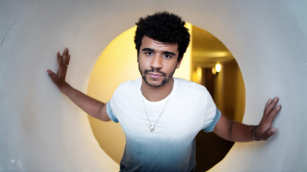 A person with curly hair wearing a white t-shirt stands inside a circular tunnel, with arms outstretched touching the sides. The background is warmly lit.