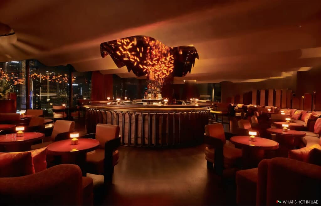 A dimly lit, elegant bar with a large circular bar counter in the center, surrounded by plush seating and small tables, with warm lighting and decorative elements hanging from the ceiling.