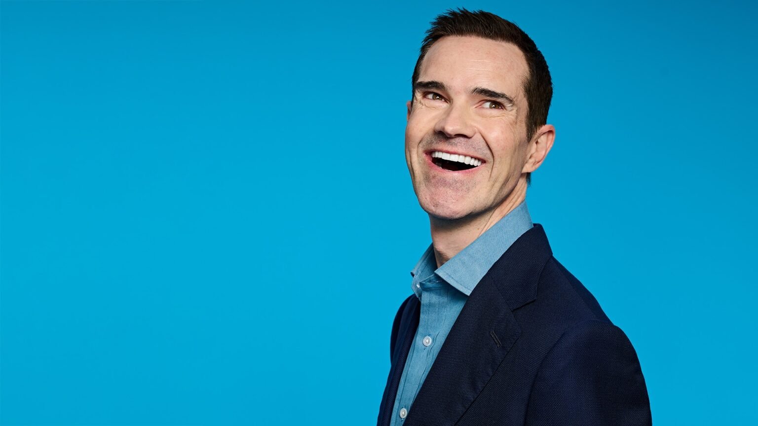 A smiling man wearing a blue shirt and dark blazer poses against a bright blue background.