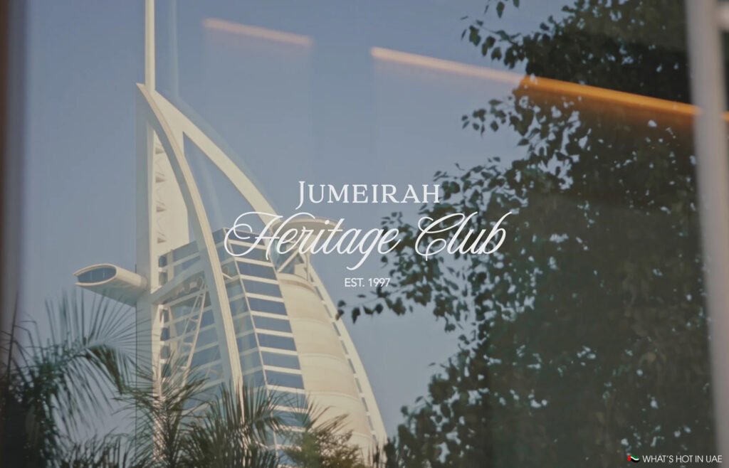 View of the Burj Al Arab hotel reflected in glass with the text 'Jumeirah Heritage Club Est. 1997' and tree branches in the foreground.