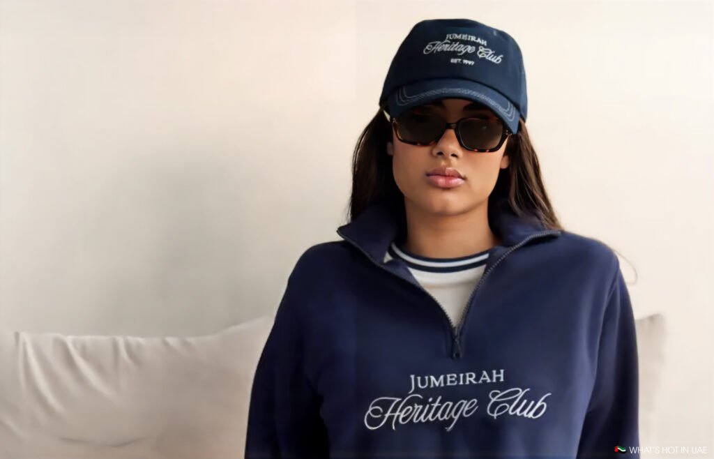 A person wearing sunglasses, a navy blue cap with "Jumeirah Heritage Club" written on it, and a matching navy blue zip-up top with the same text.