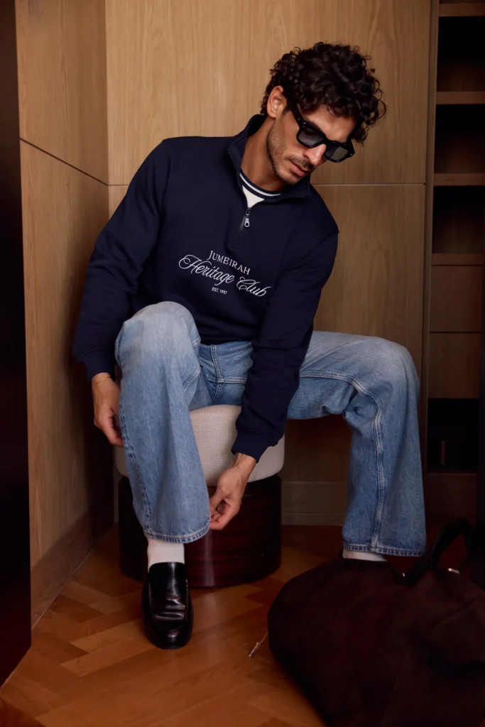 A person wearing sunglasses, a navy sweatshirt with "Jumeirah Heritage Club" text, light blue jeans, and black loafers, sitting on a stool adjusting their trousers in a modern wooden room.