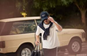A man wearing a cap, sunglasses, and a white T-shirt with a navy sweatshirt draped over his shoulders walks while holding a black bag. A vintage beige SUV is parked in the background with trees providing shade.