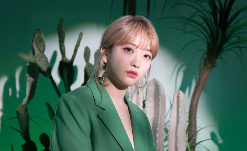 Punch, with light brown hair and hoop earrings wearing a green jacket is posing in front of a backdrop featuring cacti and plants.