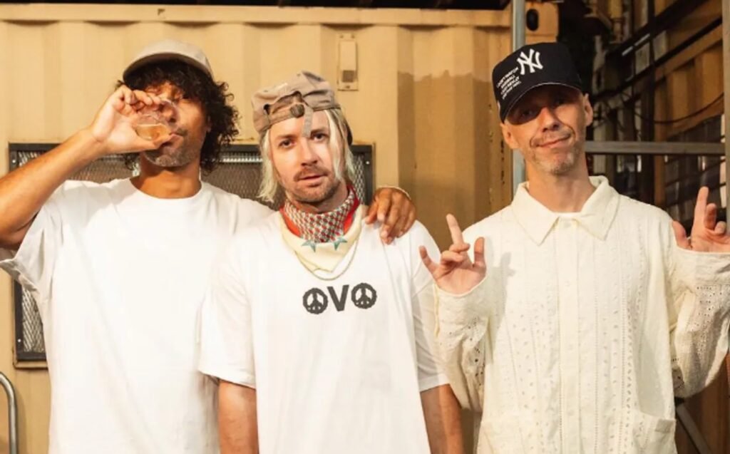 Keinemusik standing together, each wearing different casual outfits, with the man on the left holding a drink, the man in the middle wearing a cap backwards and a necklace, and the man on the right making a hand gesture.