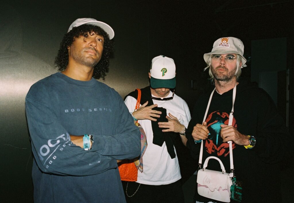 Keinemusik stand together, each wearing a hat and casual clothing, with accessories such as bags and sunglasses in a dimly lit setting.