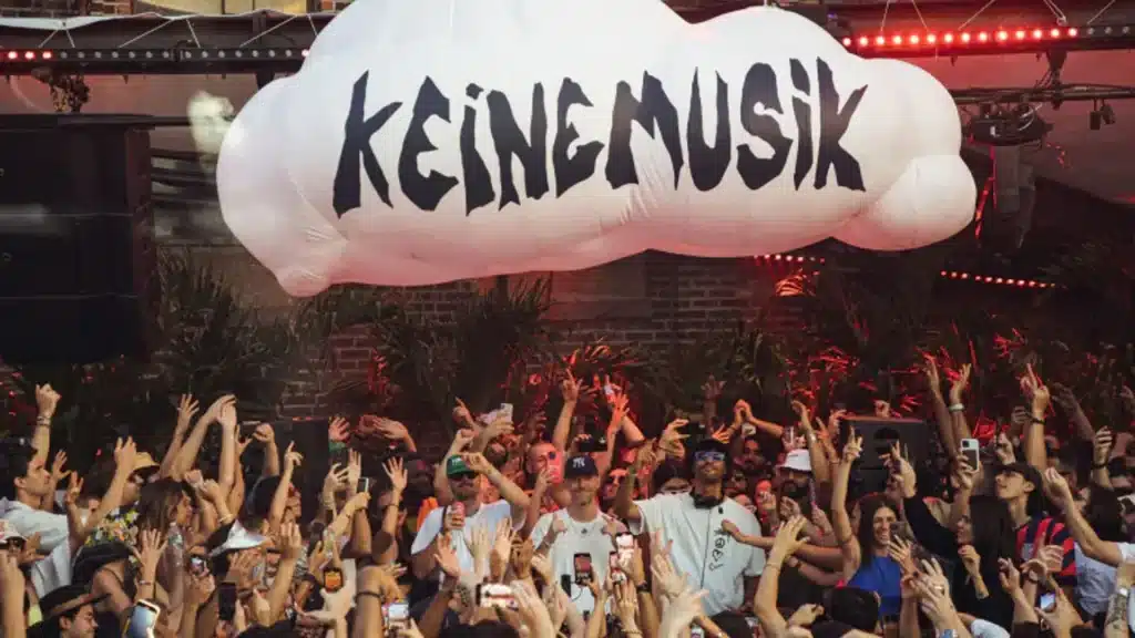 A crowd of people with raised hands at a lively event beneath a large white cloud-shaped decoration with "KEINE MUSIK" written on it.