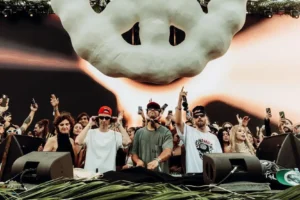 A group of people at a music event with one person in a white shirt and red cap using headphones at a DJ booth, surrounded by a crowd holding smartphones. Above them is a large, abstract cloud-like decoration.