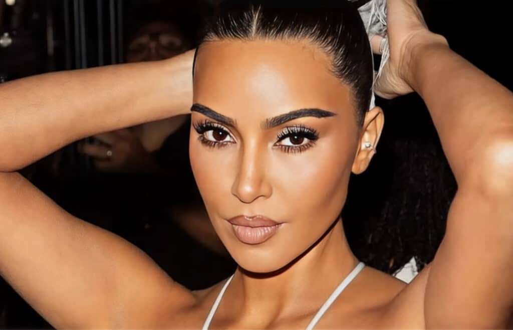 A person with long dark hair pulled back, wearing makeup, and holding their arms behind their head in a confident pose.