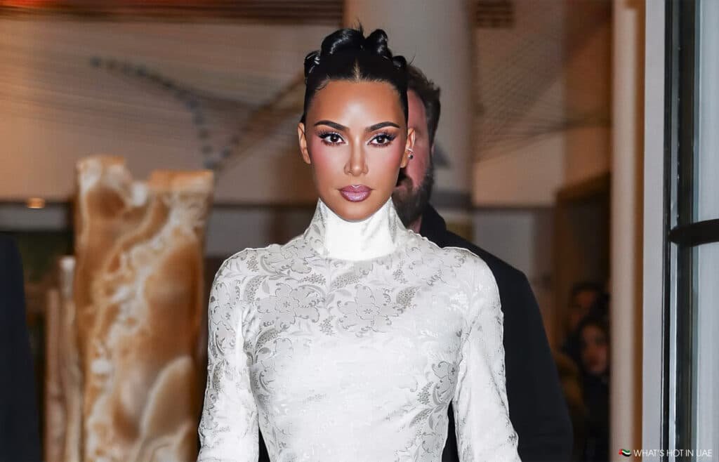Person wearing a high-necked, white floral pattern dress with styled hair, standing in an indoor setting.
