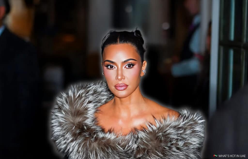 Kim Kardashian with striking makeup and a high bun hairstyle, wearing a luxurious fur coat, stands against a blurred indoor background.