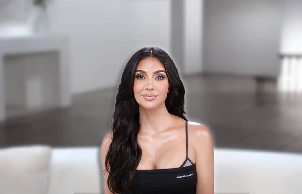 A woman with long dark hair wearing a black tank top is seated indoors against a blurred neutral background.