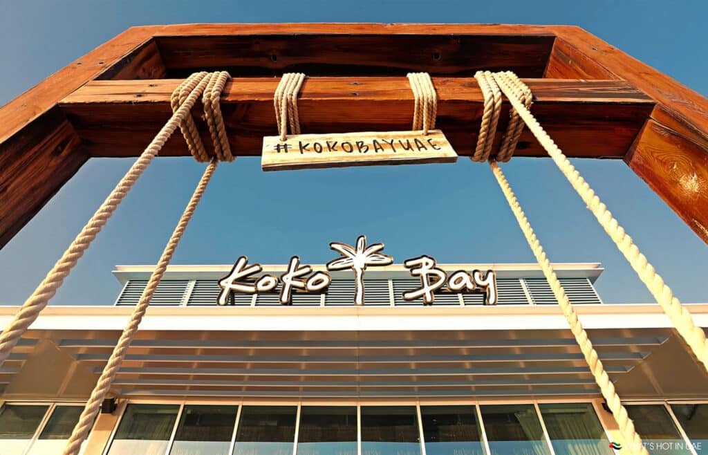Entrance of Koko Bay restaurant with a wooden frame hanging a sign that reads '#KokoBayUAE' against a clear blue sky.