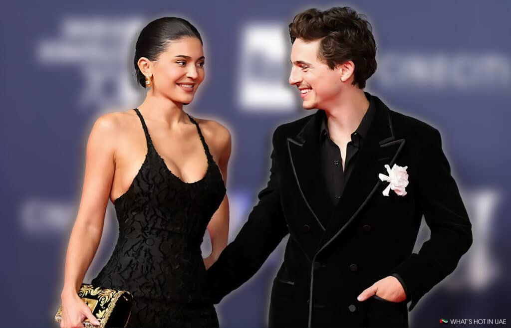 Kylie Jenner and Timothée Chalamet on a red carpet; the woman is wearing a black dress with gold earrings and holding a decorative clutch, while the man is in a dark suit with a white floral boutonniere.